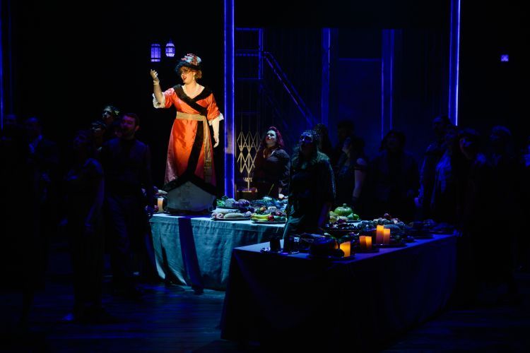 Cast of Die Fledermaus on stage with a singer in the spotlight wearing a silk red with black fur trim kimono style dress, gold sash, long gold gloves, and black ornate hat with coloured ruffles and lace veil slightly over the face